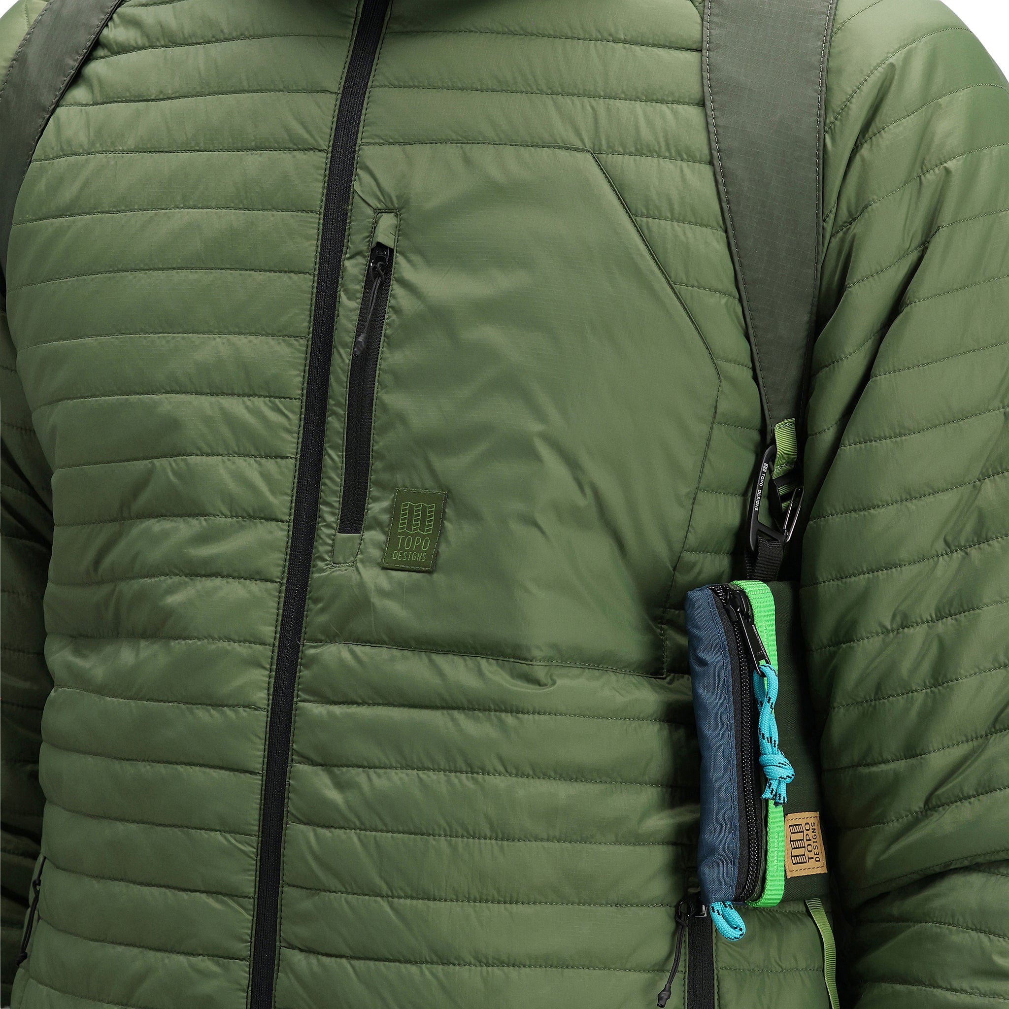 Detailaufnahme der Brusttasche und des Logoaufnähers am Model, das die Topo Designs Herren Global Puffer packbare, recycelte und isolierte Hoodie-Jacke in "olive" grün trägt.